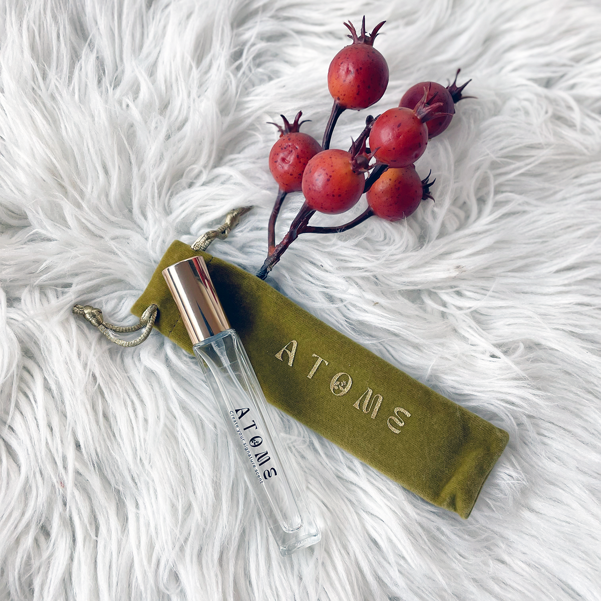 A clear glass perfume sample vial with a gold cap lies on a white faux-fur surface beside an olive green drawstring pouch embossed with the brand name ATOME. A branch with red berries is placed above the items as styling props.