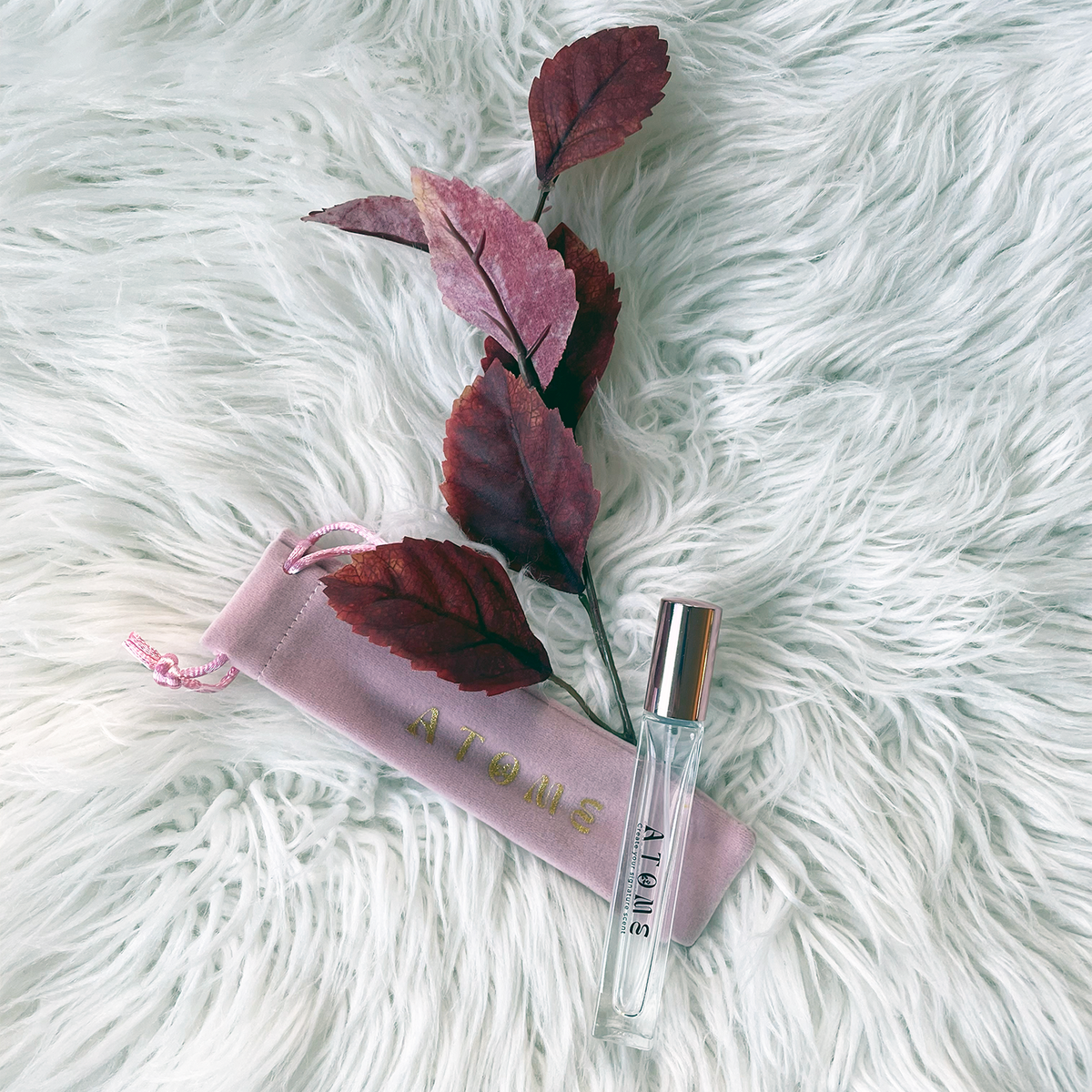 A clear glass perfume sample vial with a silver cap lies on a white faux-fur surface beside a pale pink drawstring pouch printed with the brand name 'ATOME'. A decorative stem with burgundy leaves is placed between the vial and pouch.