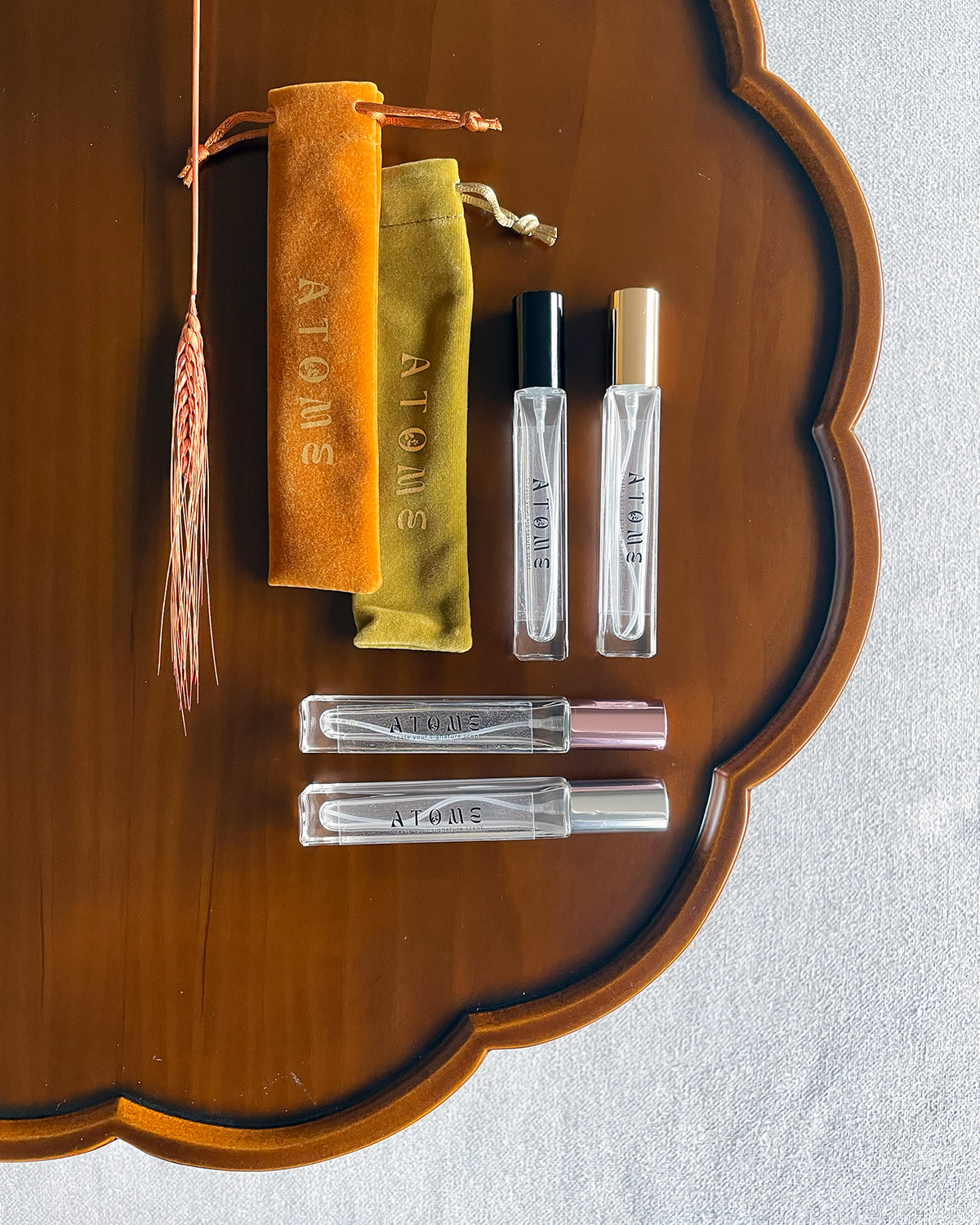Four small perfume sample vials and two matching fabric pouches from the brand ATOME, arranged on a scalloped wooden tray with a dried wheat stem prop.