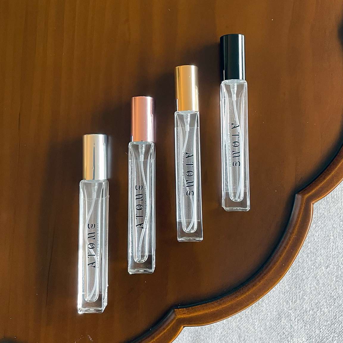 Four clear glass perfume roller bottles with silver, rose gold, gold, and black caps lined up on a scalloped wooden tray.